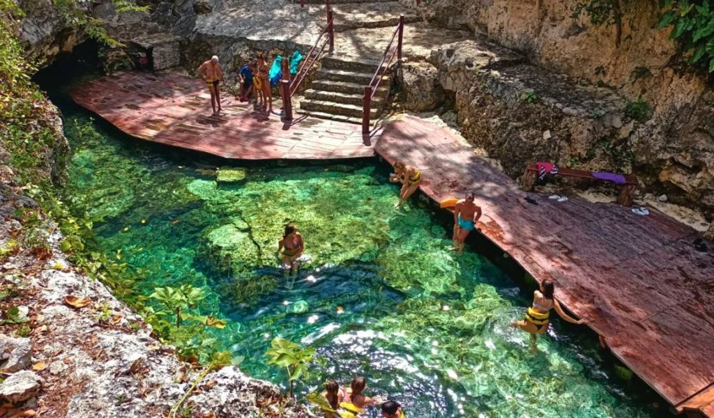 바위로 둘러싸인 맑고 청록색의 천연 수영장과 계단이 있는 나무 데크에서 수영하고 휴식을 취하는 사람들.