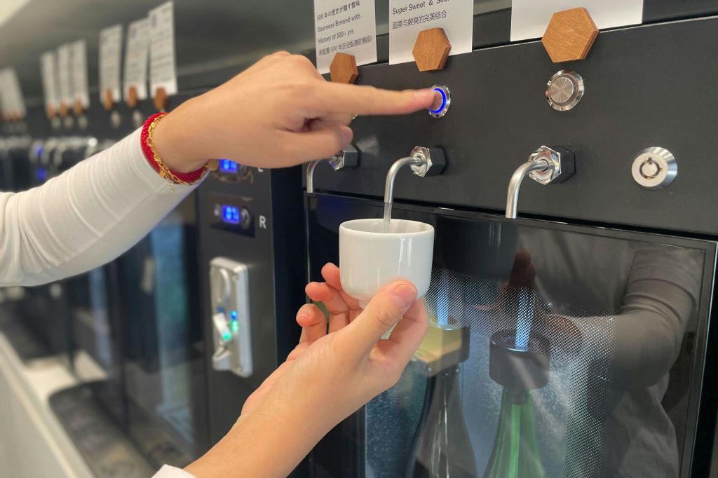 사람이 파란색 점등 버튼을 눌러 음료를 기기의 흰색 컵에 넣습니다. 플라스틱 병은 아래에서 부분적으로 볼 수 있습니다.