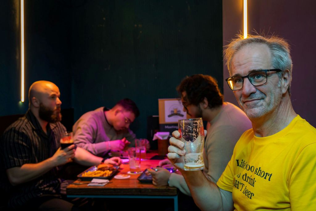 노란 셔츠를 입은 남성이 조명이 어두운 방에서 유리를 들고 있으며, 사람들이 테이블에 앉아 있습니다.