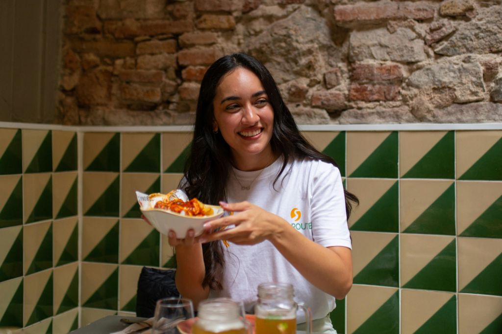 미소를 지으며 음식 접시를 들고 있는 여성이 테이블에 앉아 음료를 마시고 있습니다. 배경에는 벽돌 벽과 녹색 및 베이지색 타일이 있습니다.