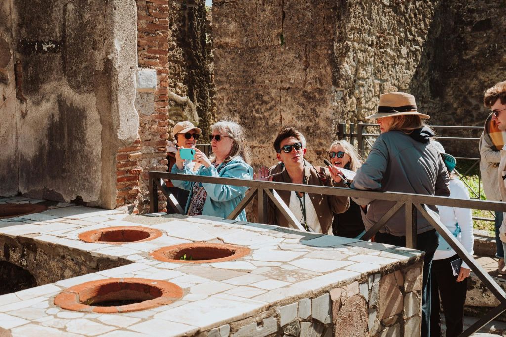 고대 유적 근처에 서서 사진을 찍고 있는 한 무리의 사람들로, 전경에 타일로 마감된 구조물이 있습니다.