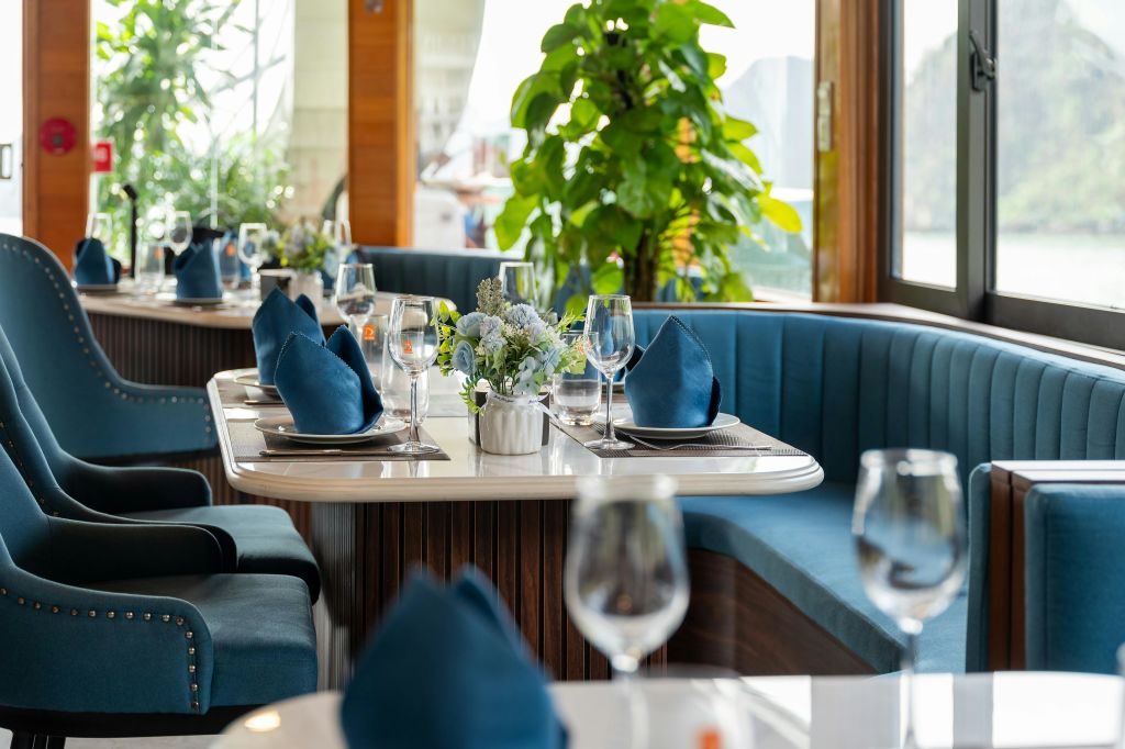푸른색 냅킨과 유리 공예품, 꽃무늬 센터피스가 놓인 레스토랑 테이블로, 쿠션이 있는 좌석과 화분을 배경으로 하고 있습니다.