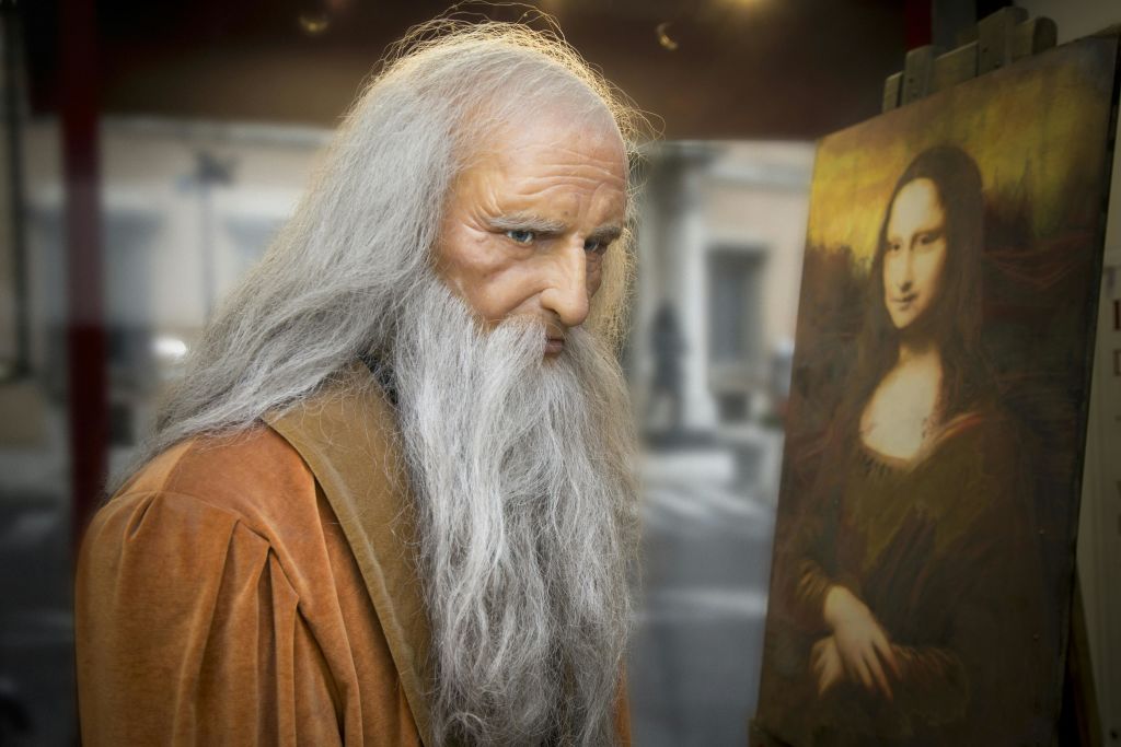 긴 흰 머리에 수염을 기른 사람이 실내 이젤에 그려진 모나리자 그림 옆에 서 있습니다.