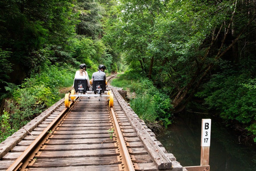 페달을 밟은 두 사람이 작은 개울 옆 숲이 우거진 철로를 따라 달립니다.