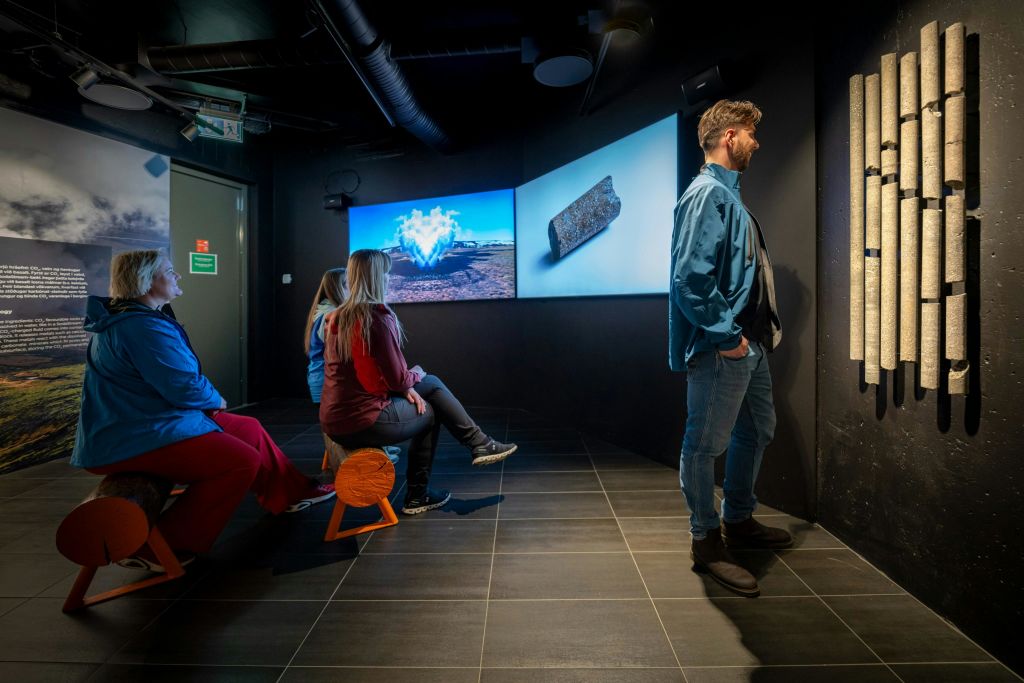 조명이 희미한 방에 있는 사람들은 현대적인 인테리어와 좌석이 있는 풍경과 사물의 이미지를 보여주는 대형 스크린을 보고 있습니다.