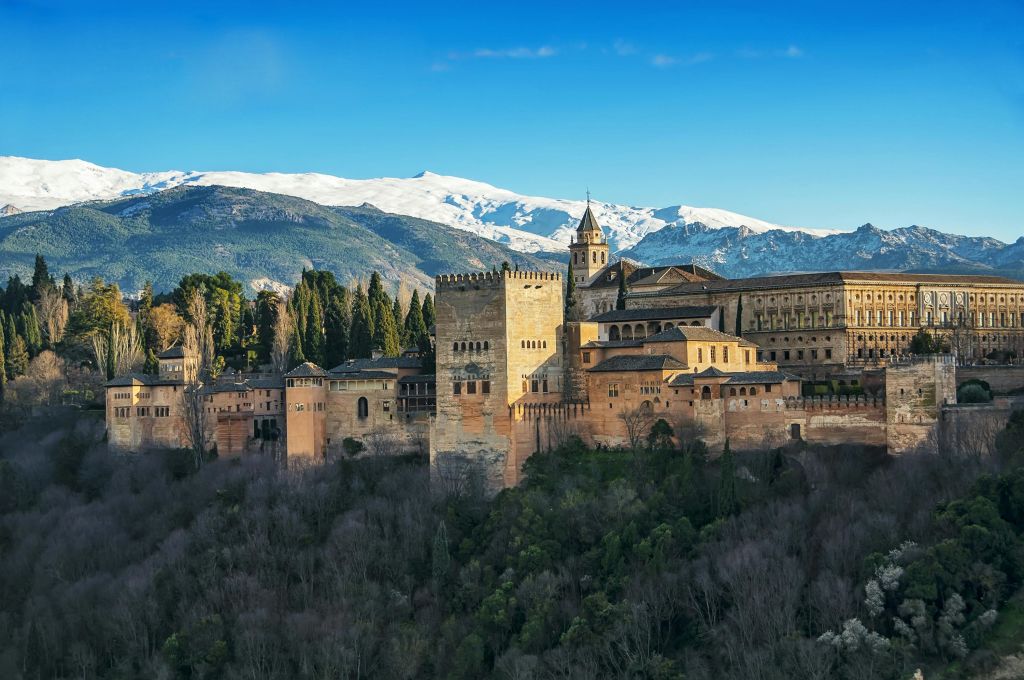 눈 덮인 산과 맑고 푸른 하늘을 배경으로 나무가 우거진 언덕에 탑과 건물이 있는 역사적인 요새입니다.