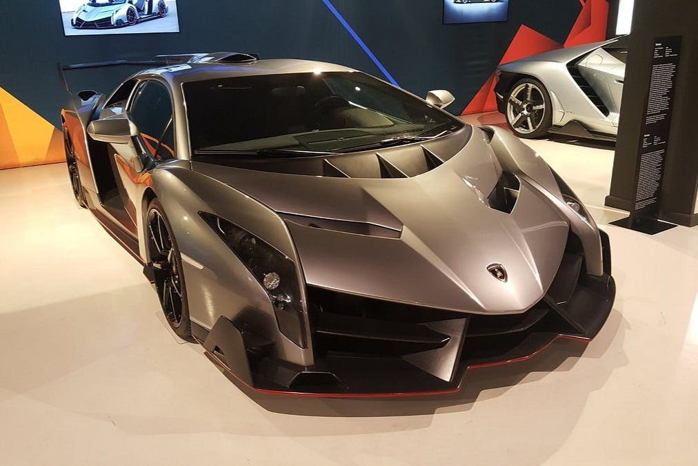 각도 디자인이 돋보이는 세련된 메탈릭 그레이 스포츠카가 쇼룸에 전시되어 있으며, 다른 차량이 배경에 보입니다.