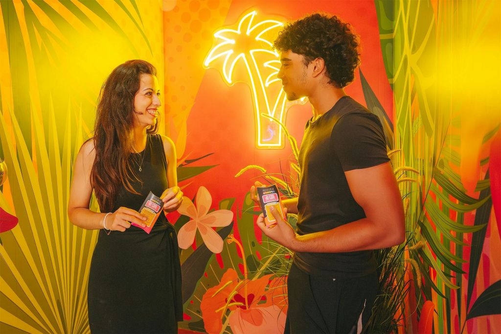 한 여성과 한 남성이 소포를 들고 서로에게 미소를 짓고 있습니다. 그들은 밝고 열대 테마의 배경 앞에 서 있습니다.
