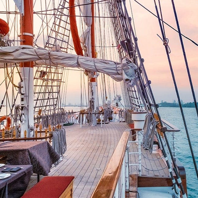 석양을 감상할 수 있는 돛단배의 나무 데크, 리깅, 밧줄, 돛. 왼쪽에 식탁보와 의자가 놓인 테이블이 보입니다.