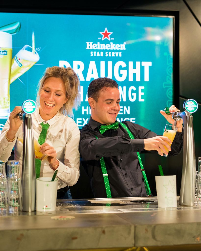 하이네켄 브랜딩을 배경으로 두 사람이 바에서 미소를 지으며 맥주를 붓고 있습니다.