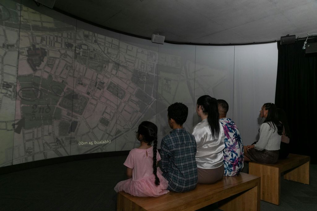 나무 벤치에 앉아 조명이 어두운 방의 곡선형 화면에서 커다란 지도 투영을 보고 있는 사람들이 여러 명 있습니다.