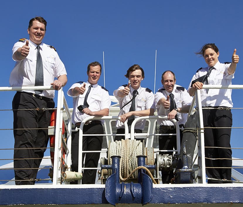 파란 하늘을 배경으로 난간 뒤에 서서 엄지손가락을 치켜세우는 선박에 제복을 입은 승무원 5명이 타고 있습니다.
