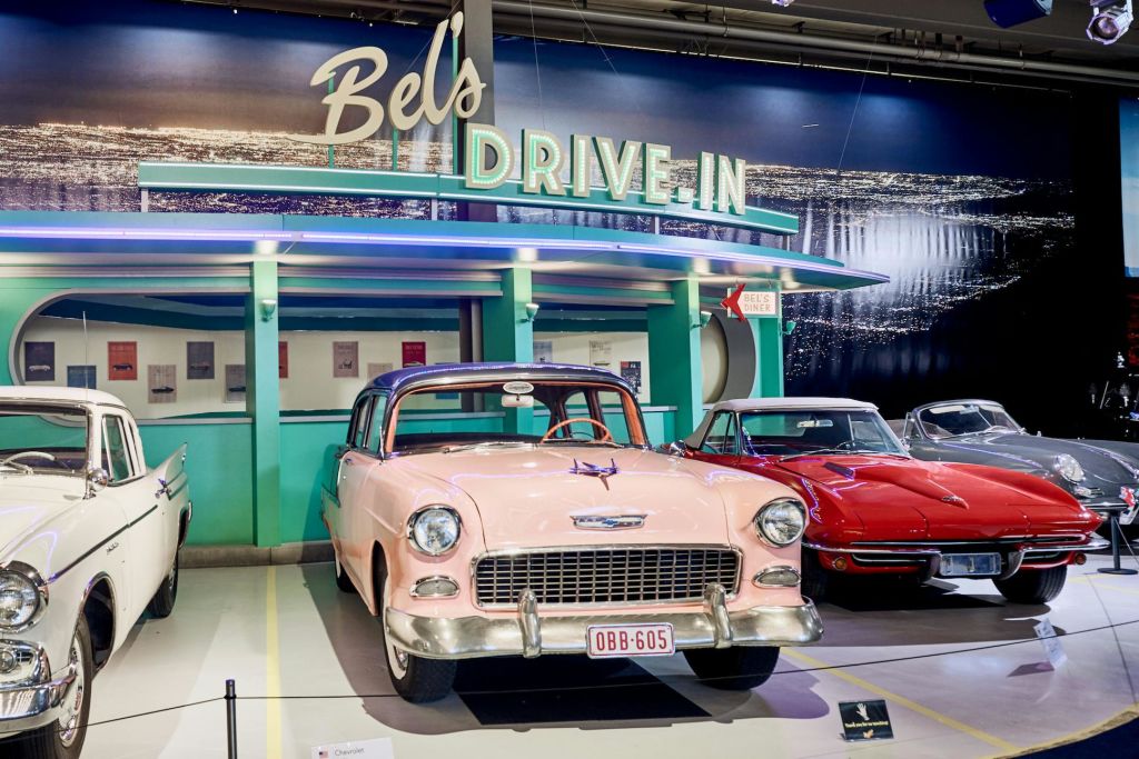 네온 사이니지와 녹색 및 파란색 외관이 있는 복고풍 테마의 "Bel's Drive-In" 앞에 주차된 클래식한 핑크색 차량.