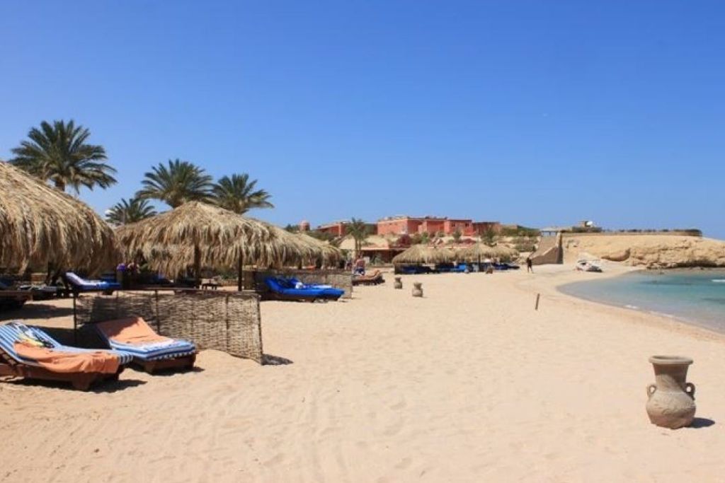 맑고 푸른 하늘 아래 야자수와 건물을 배경으로 한 모래 해변의 일광욕용 파라솔과 라운지 의자.