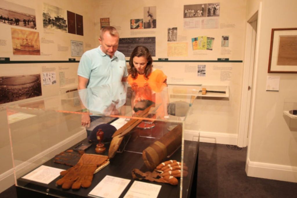 두 사람이 박물관 전시물에 전시된 장갑, 박쥐, 문서 등 역사적인 스포츠 기념품을 관찰합니다.