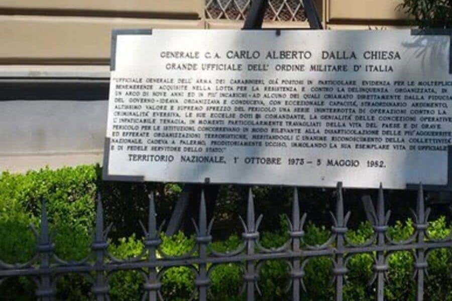 녹색 단풍이 있는 검은 철제 울타리 뒤에 제네랄 C.A. 카를로 알베르토 달라 키에사를 기리는 금속 상패가 전시되어 있습니다.