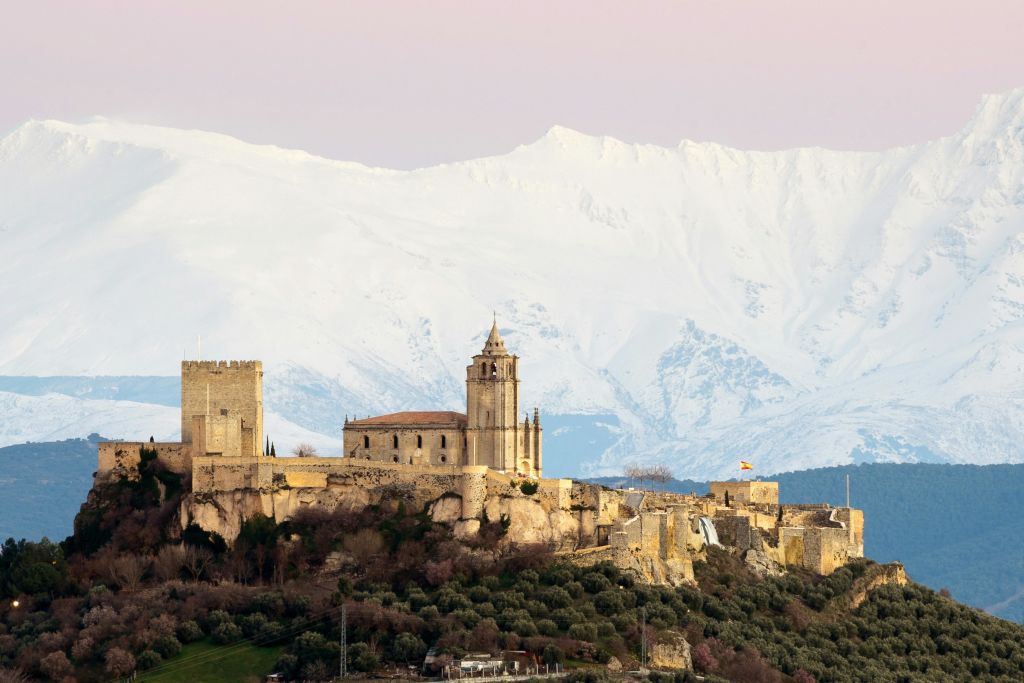 분홍빛 하늘 아래 눈 덮인 산을 배경으로 언덕 꼭대기에 있는 중세 성곽과 교회.