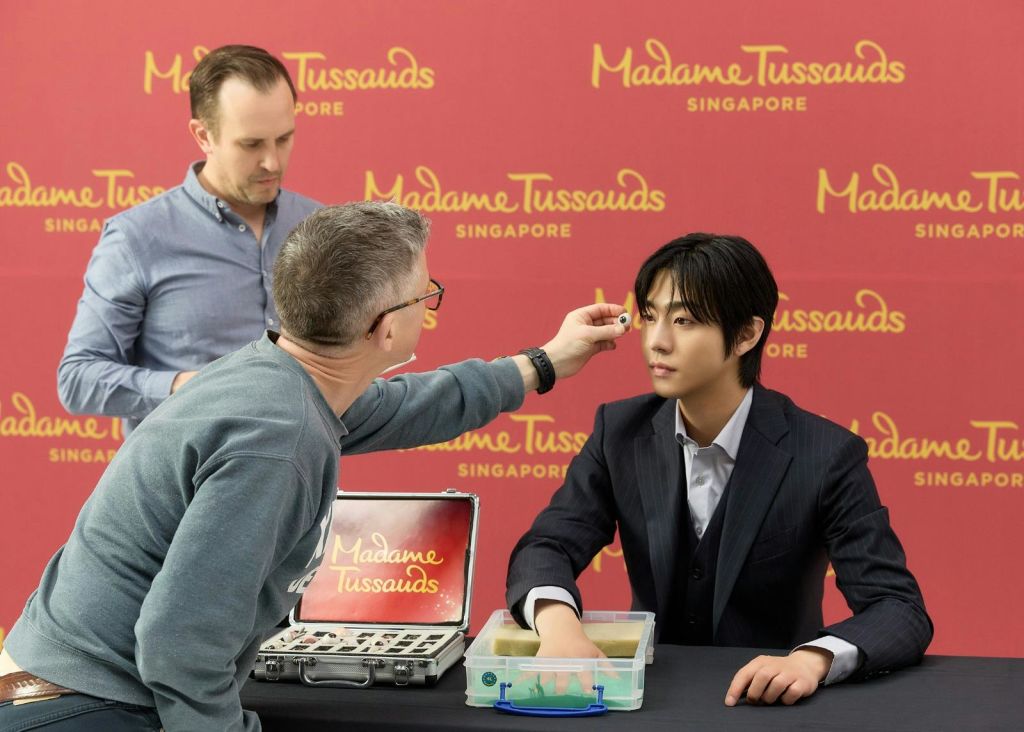 한 사람이 싱가포르 마담 투소 (Madame Tussauds Singapore) 의 왁스 피규어의 디테일을 조정하고 있으며, 도구와 재료가 보입니다.