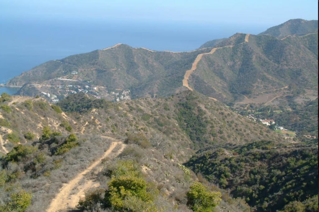 희박한 초목, 먼 산, 집, 바다를 배경으로 건조하고 언덕이 많은 지형을 구불구불한 하이킹 트레일이 펼쳐집니다.
