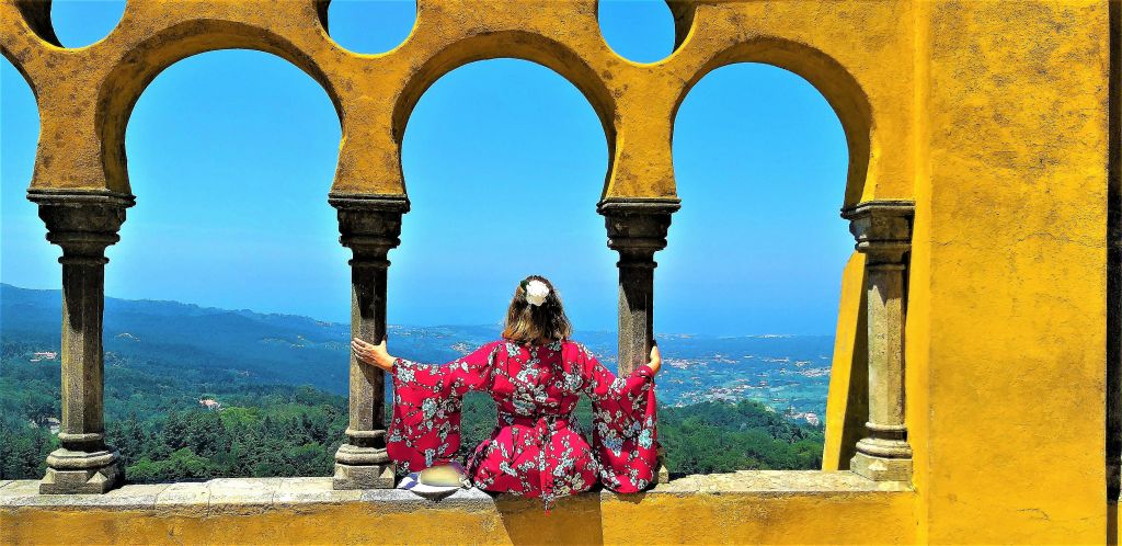 노란색 아치 사이에 앉아 언덕, 녹지, 푸른 하늘이 있는 경치 좋은 풍경을 내려다보는 빨간색 꽃무늬 드레스를 입은 사람.