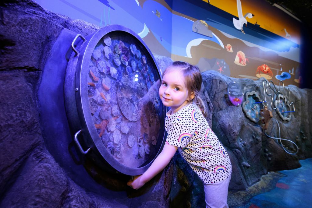 다채로운 바다 생물 벽화를 배경으로 한 어린 소녀가 둥근 수족관을 만지며 미소를 짓고 있습니다.