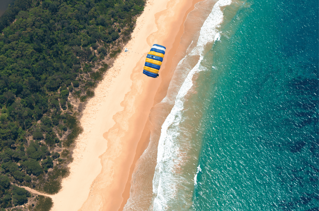 모래 해변과 청록색 바다 위로 노란색과 파란색 낙하산을 들고 패러세일링하는 사람의 공중 풍경.