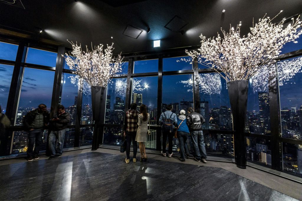 커다란 창가에 서서 밤에 도시의 스카이라인을 바라보는 사람들. 양쪽에 흰색 꽃이 달린 커다란 꽃병이 있습니다.