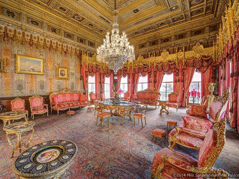 빨간색과 금색 가구, 대형 샹들리에, 정교하게 장식된 벽과 천장, 커다란 창문으로 장식된 화려한 객실.