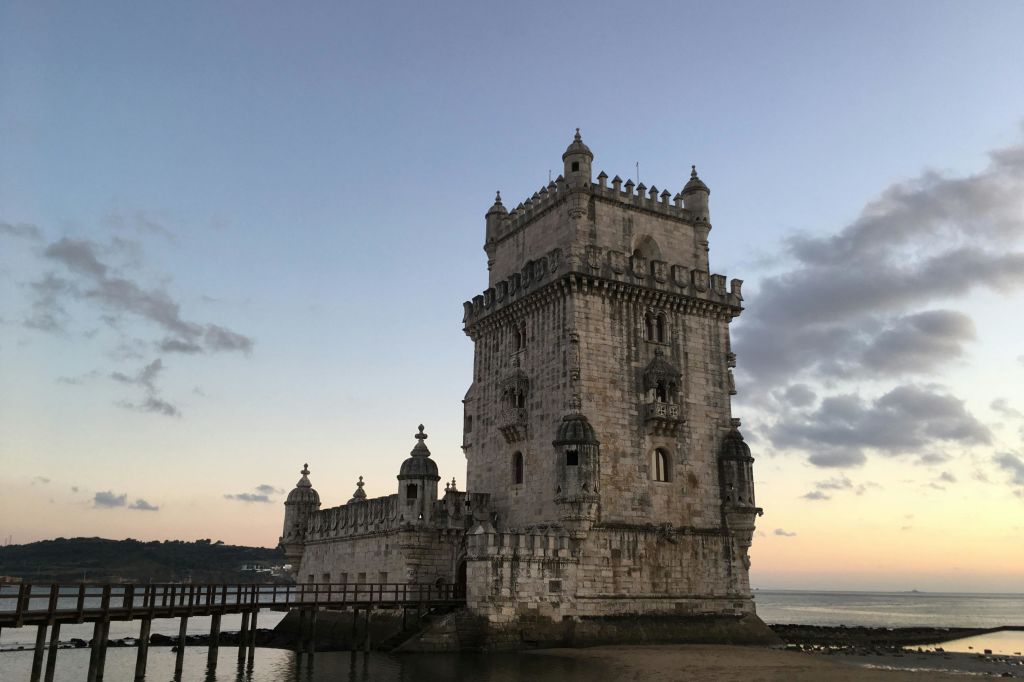 구름이 깔린 황혼의 하늘 아래, 물로 둘러싸인 전투가 있는 유서 깊은 석탑. 나무 통로가 입구로 이어집니다.