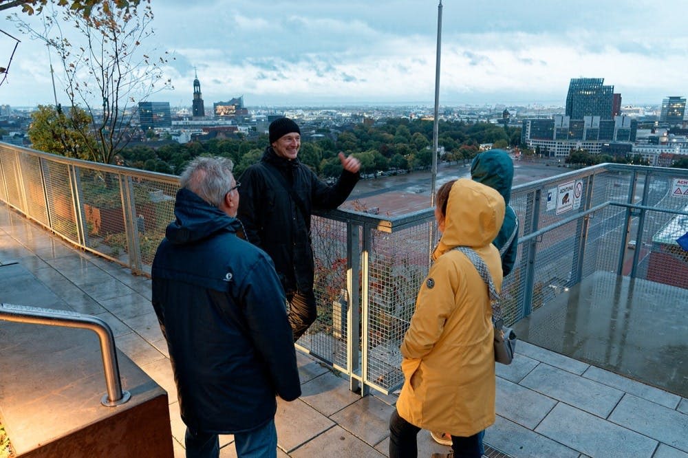 노란색 재킷을 입은 네 명이 도시가 내려다보이는 전망대에 서 있습니다. 한 사람이 말하면서 제스처를 취합니다.
