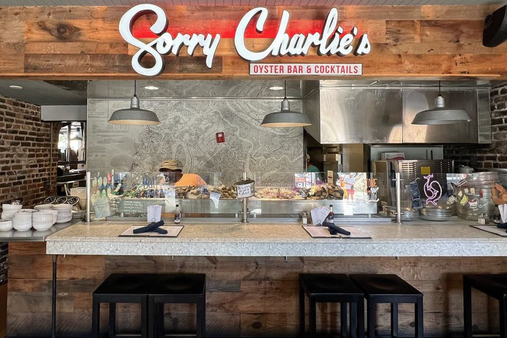 쏘리 찰리스 오이스터 바 & 칵테일 (Sorry Charlie's Oyster Bar & Cocktails) 의 카운터에는 스툴, 메뉴, 펜던트 조명 아래 해산물이 진열되어 있습니다.