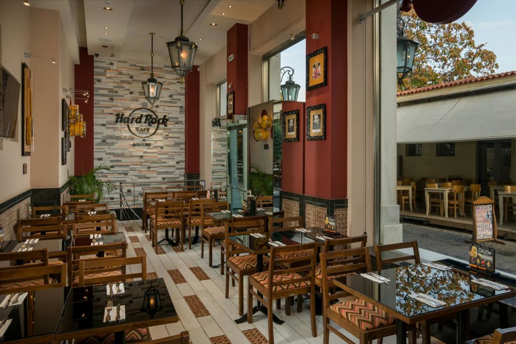 나무 테이블과 의자, 빨간색 악센트 벽, 커다란 창문, 'Hard Rock Café' 라고 적힌 간판이 있는 현대적인 레스토랑 인테리어.