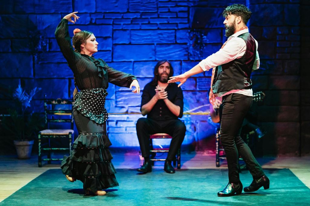 무대에서 한 여성과 한 남성이 플라멩코 댄스를 선보이고, 좌석에 앉은 뮤지션이 박수를 치고 있습니다.