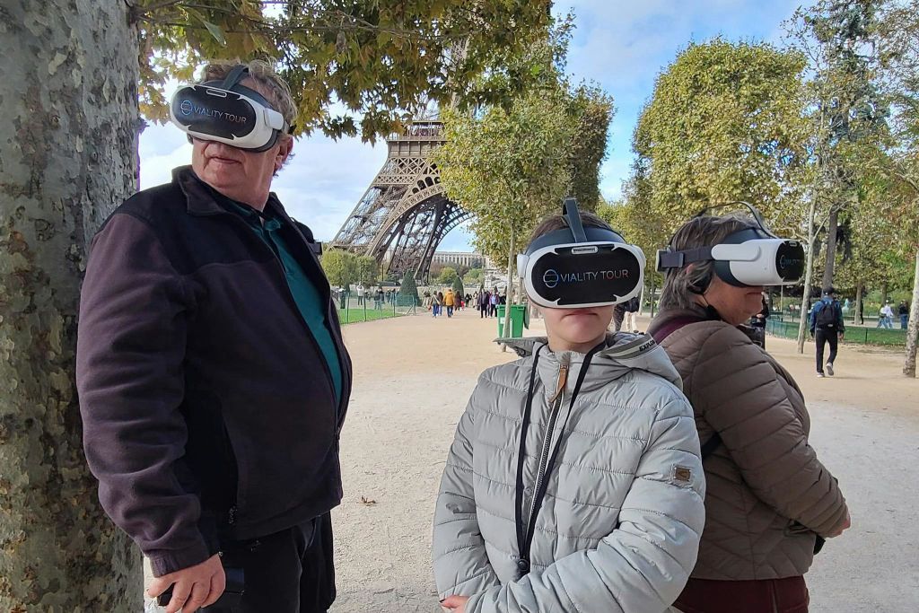 VR 헤드셋을 착용한 세 사람이 에펠탑 근처에 서서 나무와 맑은 하늘로 둘러싸여 있습니다.