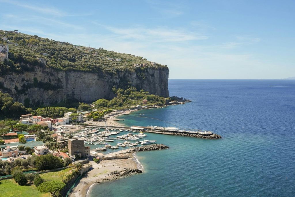 선착장, 보트, 건물, 맑은 하늘 아래 푸른 바다가 내려다보이는 절벽이 있는 해안 마을입니다.