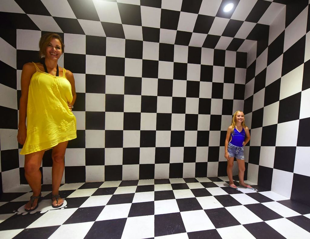 검은색과 흰색 체커보드 벽과 바닥이 있는 방에 서 있는 두 사람. 방의 관점이 크기를 왜곡합니다.