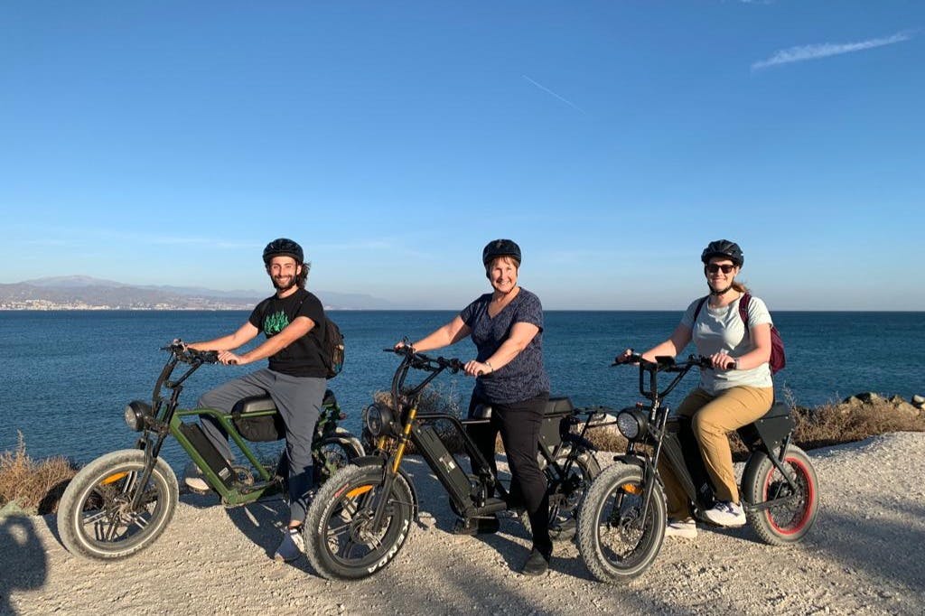 해안 도로 옆에서 전기 자전거를 타고 헬멧을 쓴 두 사람이 서 있습니다. 바다와 먼 산이 배경입니다.