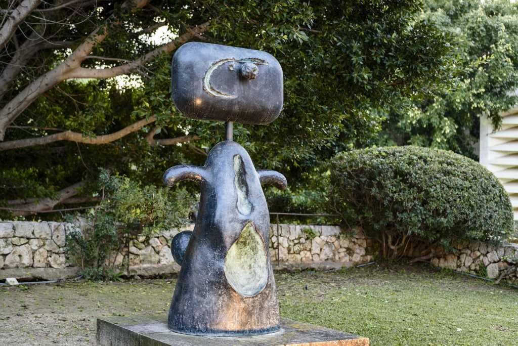 직사각형의 머리와 둥근 몸체를 가진 추상적인 청동 조각으로, 나무와 수풀 사이 야외에 세워져 있습니다.