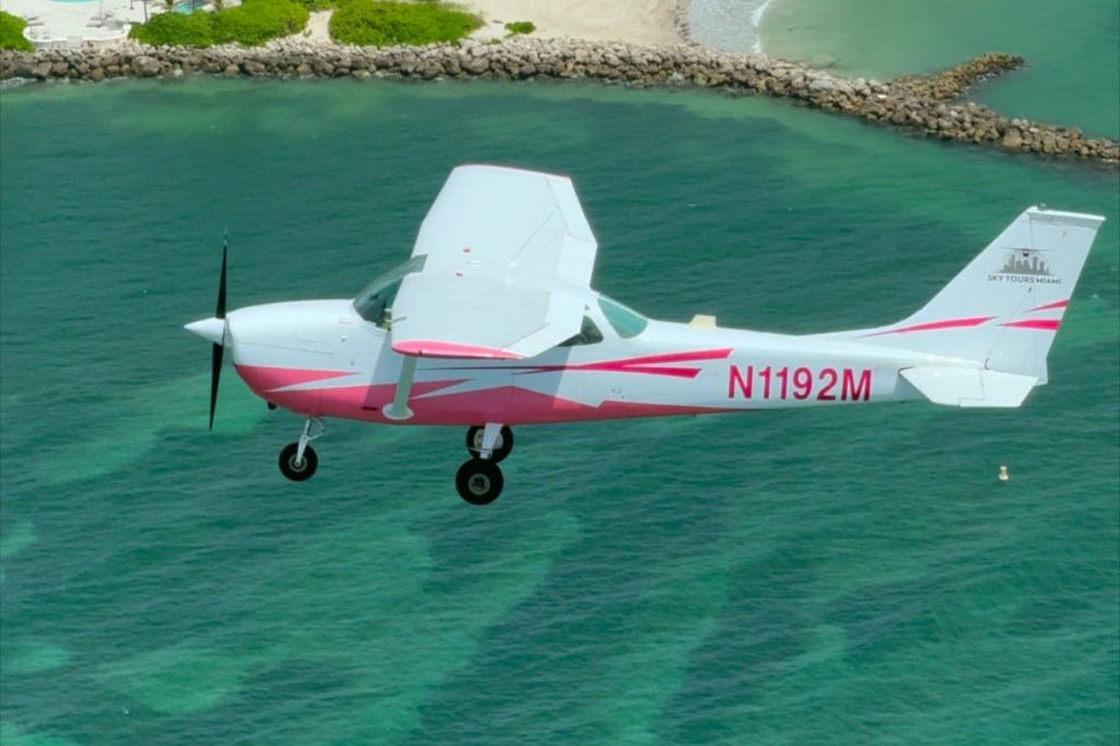 바위가 많은 해안가 근처의 맑고 푸른 바닷물 위로 날아가는 작은 분홍색과 흰색 비행기.