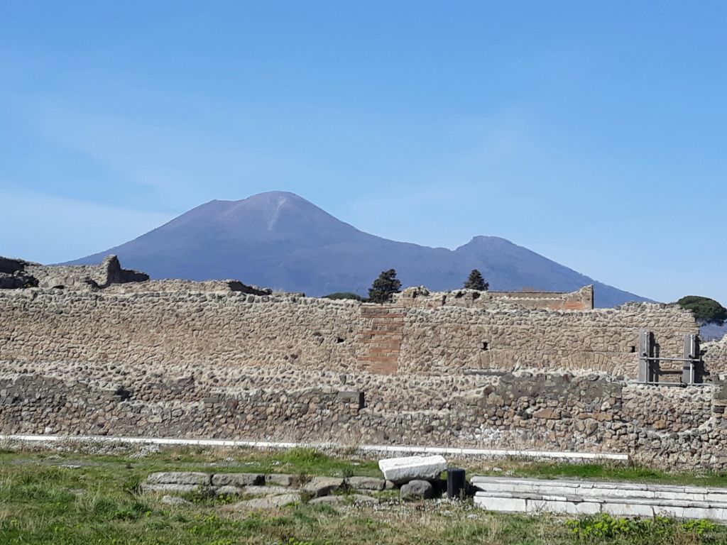 맑고 푸른 하늘 아래 잔디 전경과 베수비오 산을 배경으로 한 고대 석조 유적지.