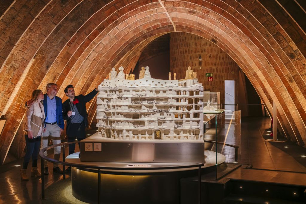 세 사람은 따뜻한 조명이 있는 아치형 벽돌 천장 아래 대형 건물의 상세한 모델을 관찰합니다.