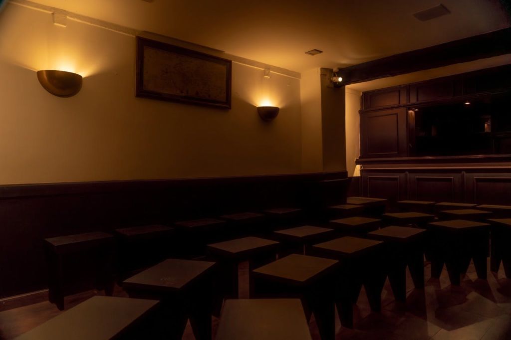 여러 개의 정사각형 스툴, 목재 패널, 벽 스콘, 벽에 액자 그림이 있는 조명이 희미한 공간입니다.