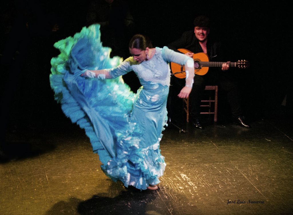 파란색 드레스를 입은 플라멩코 댄서가 활기차게 연주하는 동안 배경의 기타리스트가 연주합니다.