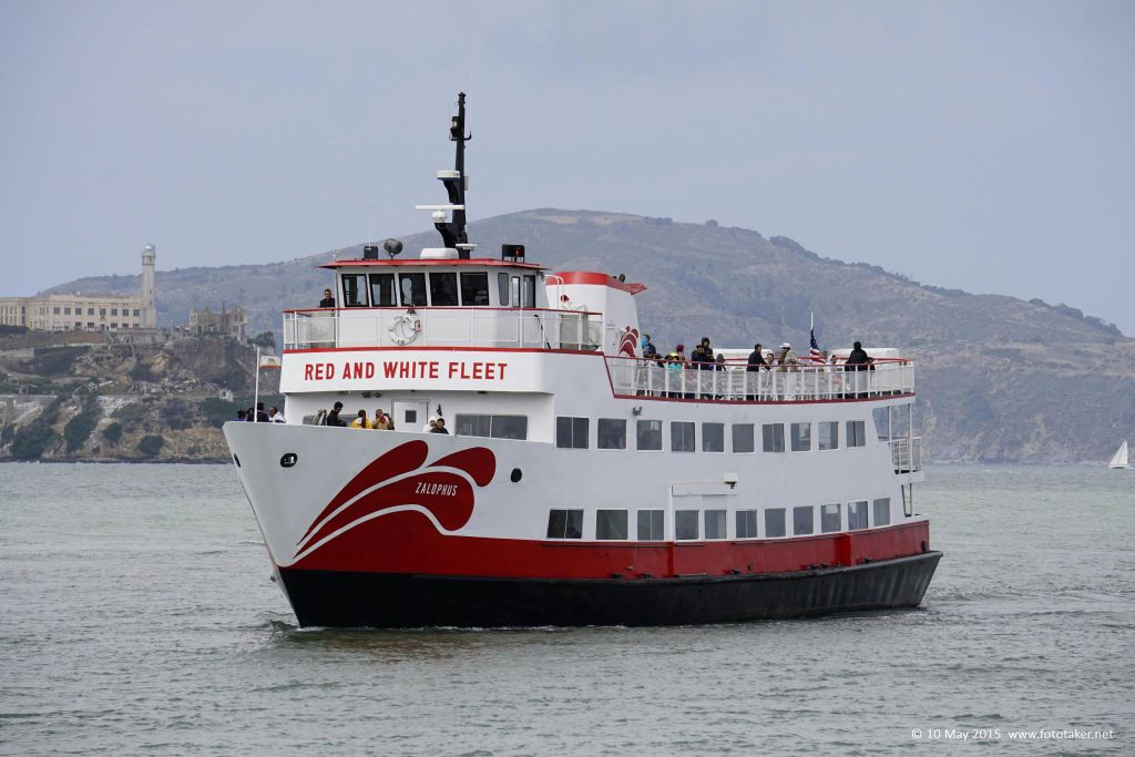 "잘로푸스" 라는 이름의 빨간색과 흰색 투어 보트에 탑승한 승객들은 언덕을 배경으로 잔잔한 물 위를 항해합니다.