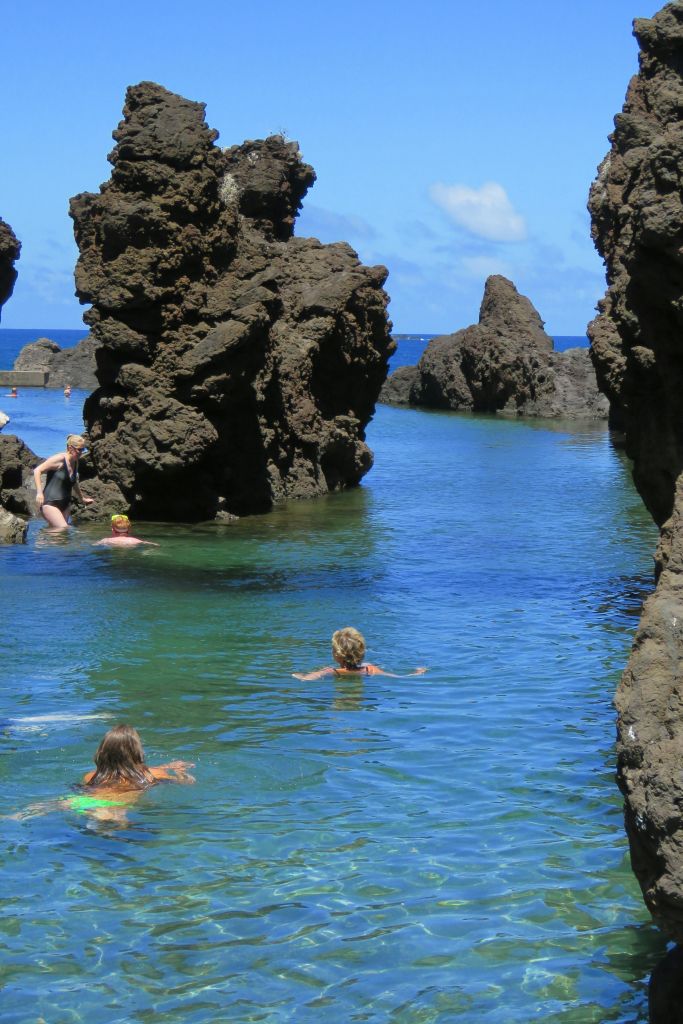 푸른 하늘 아래 커다란 암석층으로 둘러싸인 맑고 바위가 많은 조수 수영장에서 수영하는 사람들.