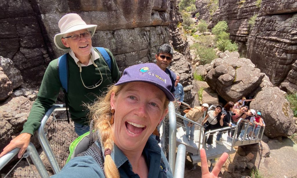 바위가 많은 등산로에서 한 무리의 사람들이 미소를 짓고 있습니다. 앞의 여성이 셀카를 찍고 다른 여성이 뒤에서 계단을 오릅니다.