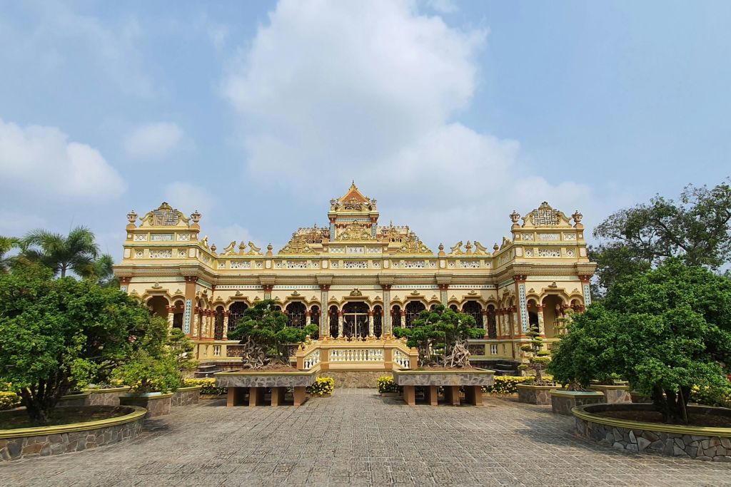부분적으로 흐린 하늘 아래 잘 손질된 식물로 둘러싸인 아치형 창문과 정교한 디테일이 돋보이는 화려한 노란색과 금색의 사원.