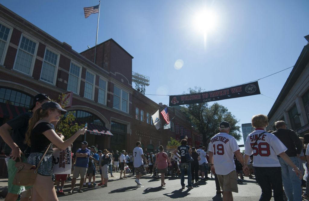 군중은 화창한 날 "Welcome to Yawkey Way" 라고 적힌 배너 아래 벽돌 경기장 밖으로 모여듭니다. 사람들은 다양한 스포츠 유니폼을 입습니다.