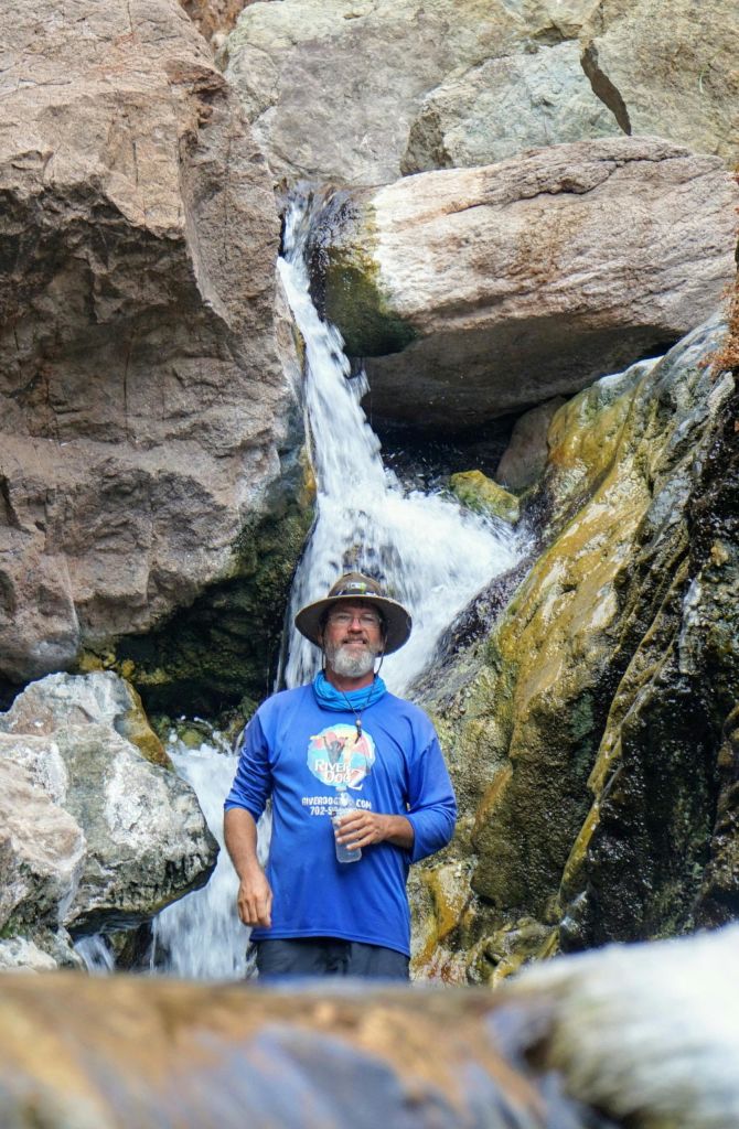 파란색 셔츠와 모자를 쓴 남성이 바위와 이끼로 둘러싸인 폭포 근처에 서 있습니다.