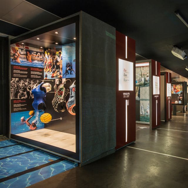 부드러운 조명 아래 벽 그래픽과 정보 패널이 있는 운동 선수의 이미지와 유니폼을 특징으로 하는 실내 스포츠 전시회입니다.